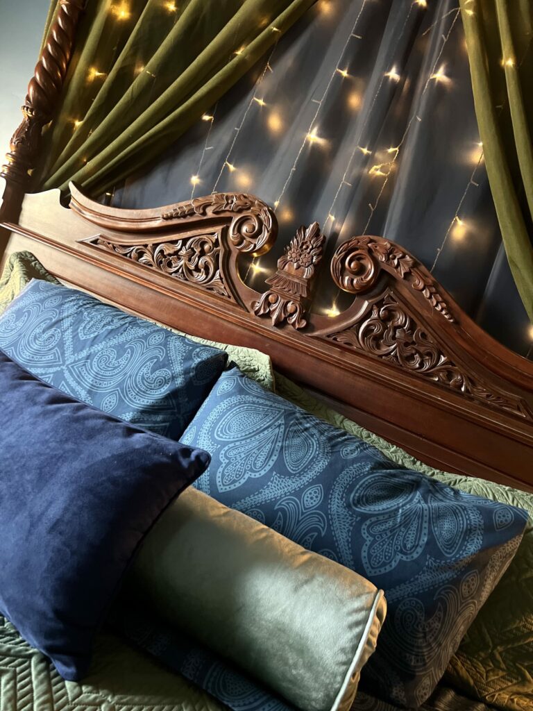 carved wood headboard with blue and green pillows and soft string lights in a wall canopy cozy bedroom setting