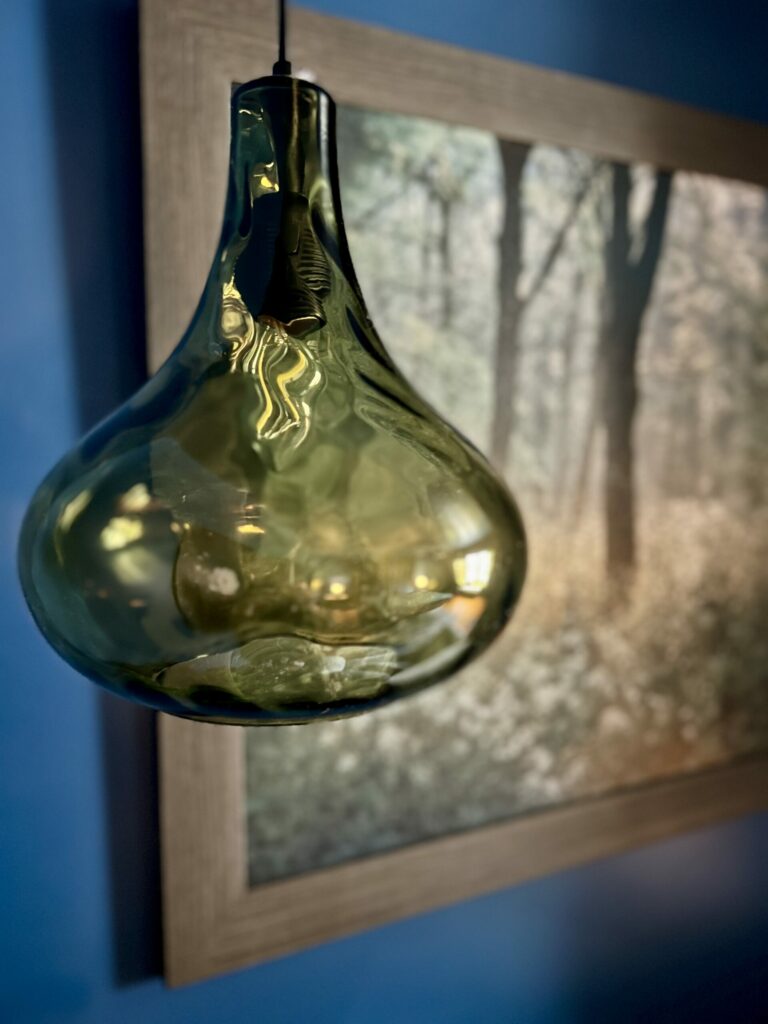 green glass pendant light glowing against blue color drenched walls and framed forest artwork in moody bedroom